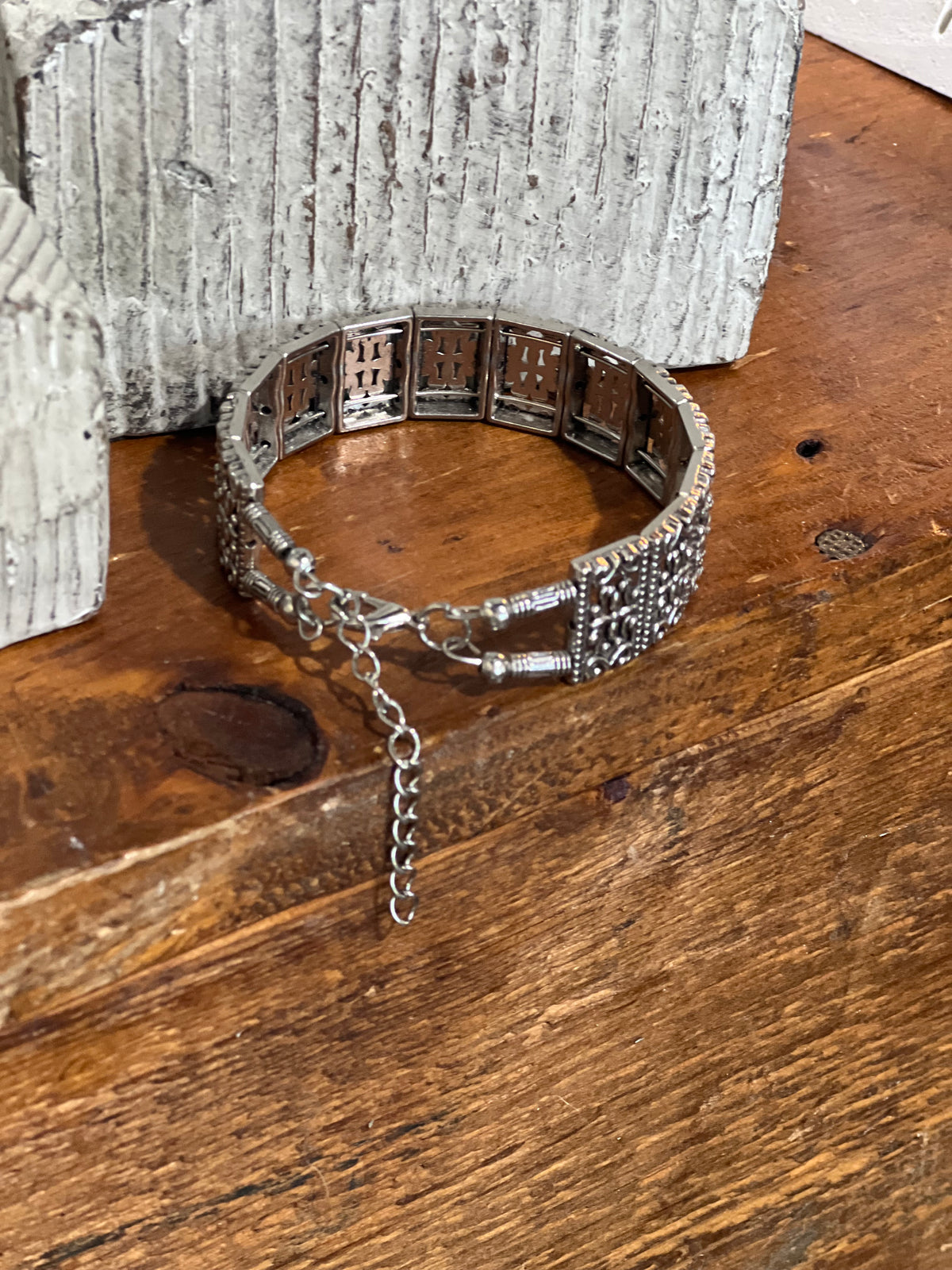 A silver-toned metal cuff bracelet with intricate cutout patterns and a dangling chain clasp, displayed on a rustic wooden surface next to a textured white stone prop.