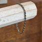 A twisted dark metal chain bracelet draped over a light wood block, set against a warm brown wooden background.