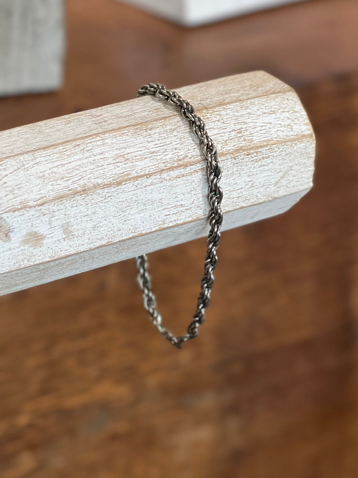 A twisted dark metal chain bracelet draped over a light wood block, set against a warm brown wooden background.
