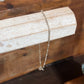 A delicate silver beaded chain necklace with a small bead pendant, draped over a light-colored wooden display block against a warm brown wooden background.