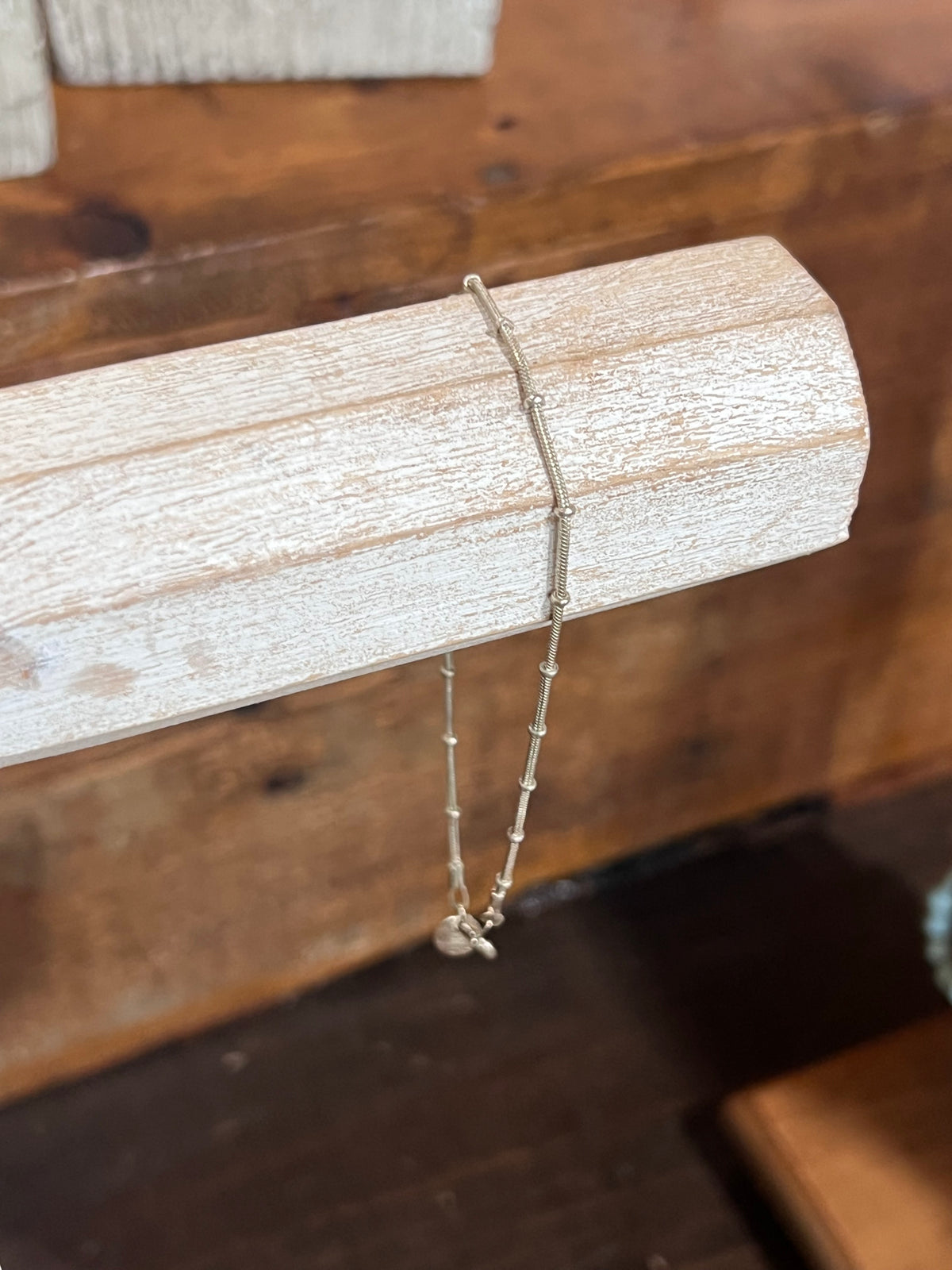 A delicate silver beaded chain necklace with a small bead pendant, draped over a light-colored wooden display block against a warm brown wooden background.