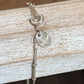 Close-up of a silver-toned chain bracelet with a lobster clasp, ring, oval charm, and a small round tag resting on a distressed white wooden surface.
