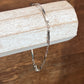 A delicate silver chain bracelet draped over a rustic white wooden block, set against a warm brown wooden tabletop background.