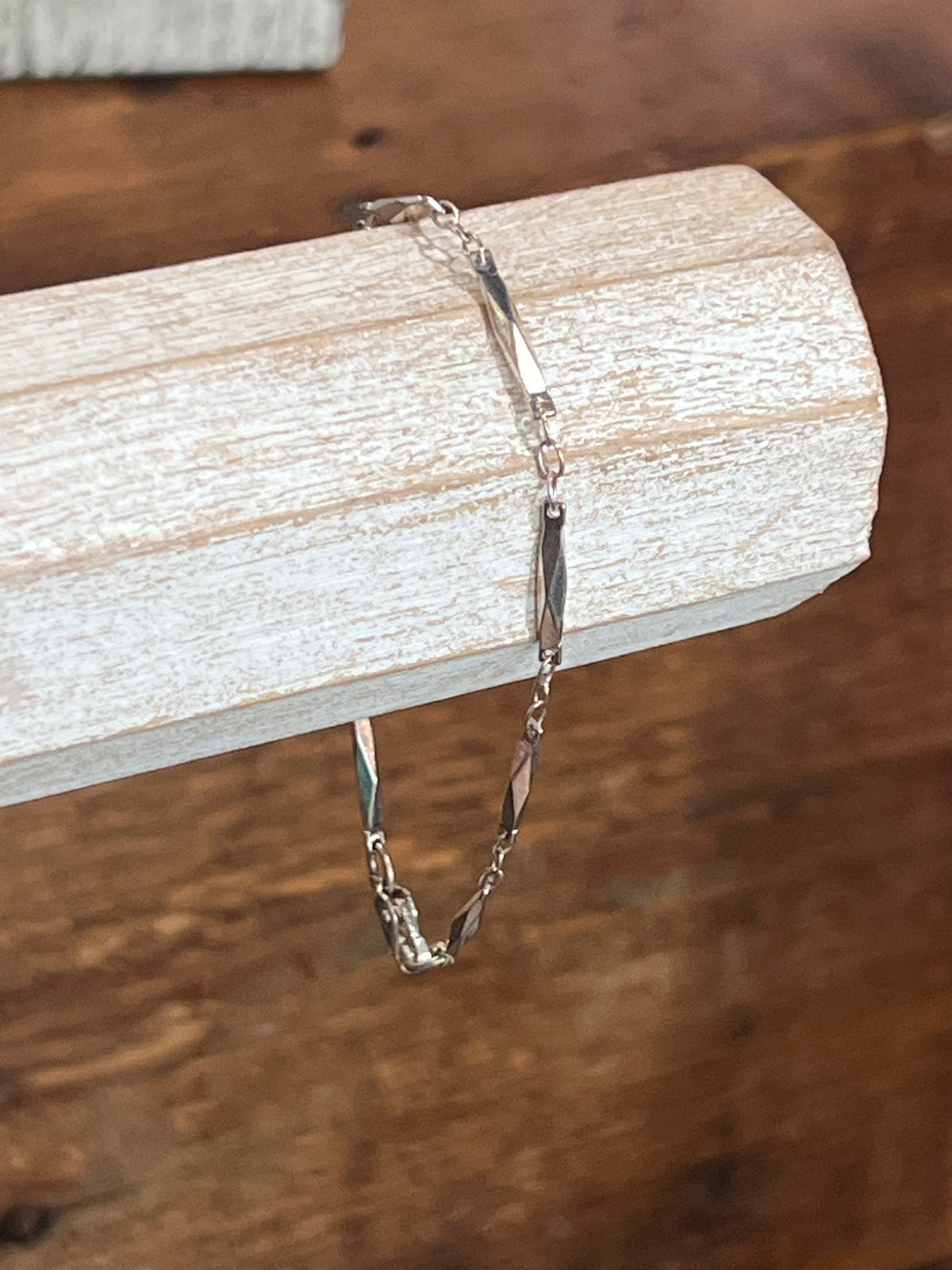 A delicate silver chain bracelet draped over a rustic white wooden block, set against a warm brown wooden tabletop background.