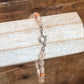 A beaded bracelet with translucent pale pink beads, small orange accent beads, and a silver clasp, displayed on a white wood block against a rustic wooden background.