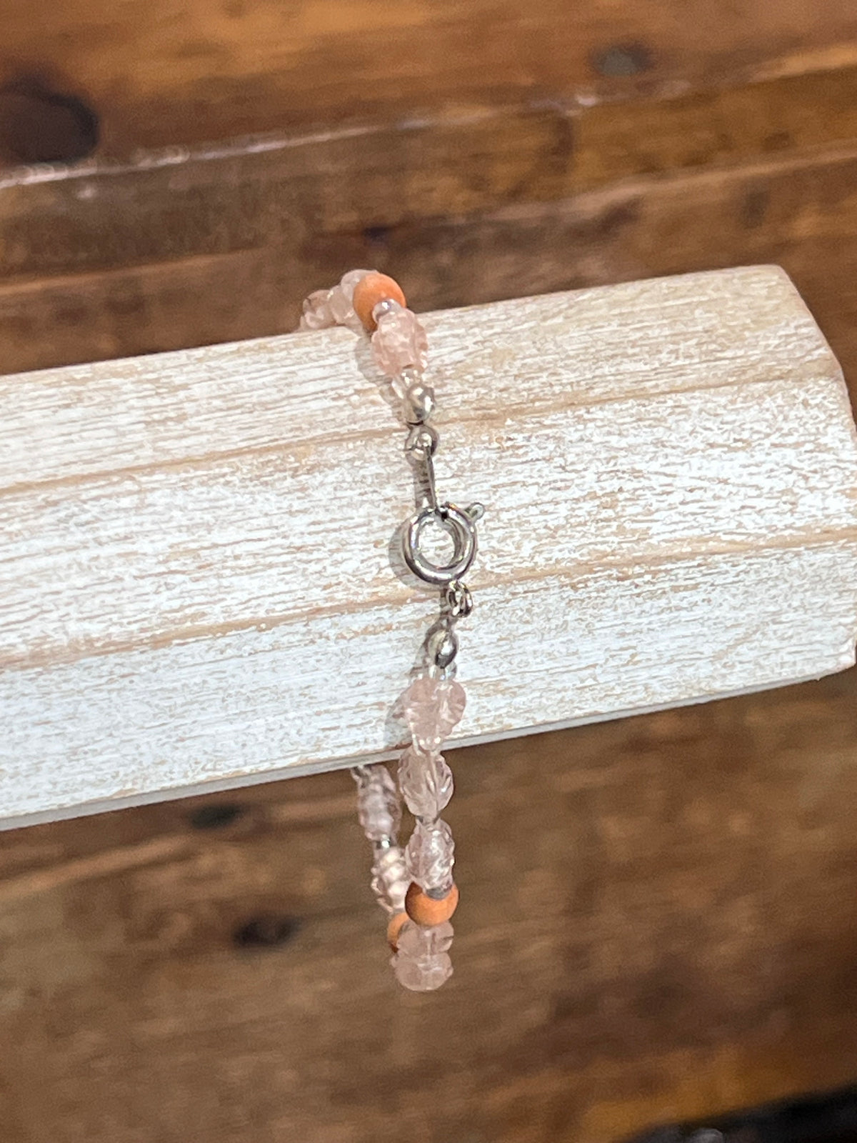A beaded bracelet with translucent pale pink beads, small orange accent beads, and a silver clasp, displayed on a white wood block against a rustic wooden background.