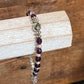 A beaded stretch bracelet with dark purple faceted beads, silver-toned textured beads, and a central iridescent green bead, displayed on a light wood block over a rustic wooden background.