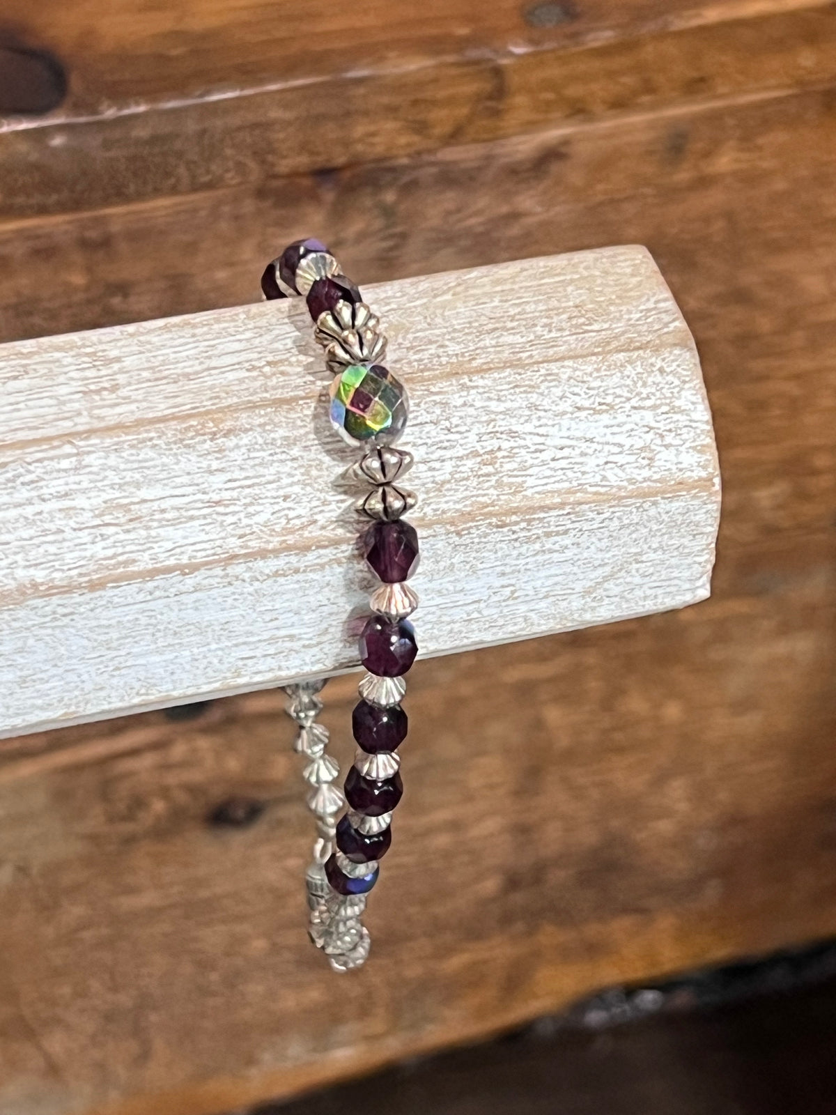 A beaded stretch bracelet with dark purple faceted beads, silver-toned textured beads, and a central iridescent green bead, displayed on a light wood block over a rustic wooden background.