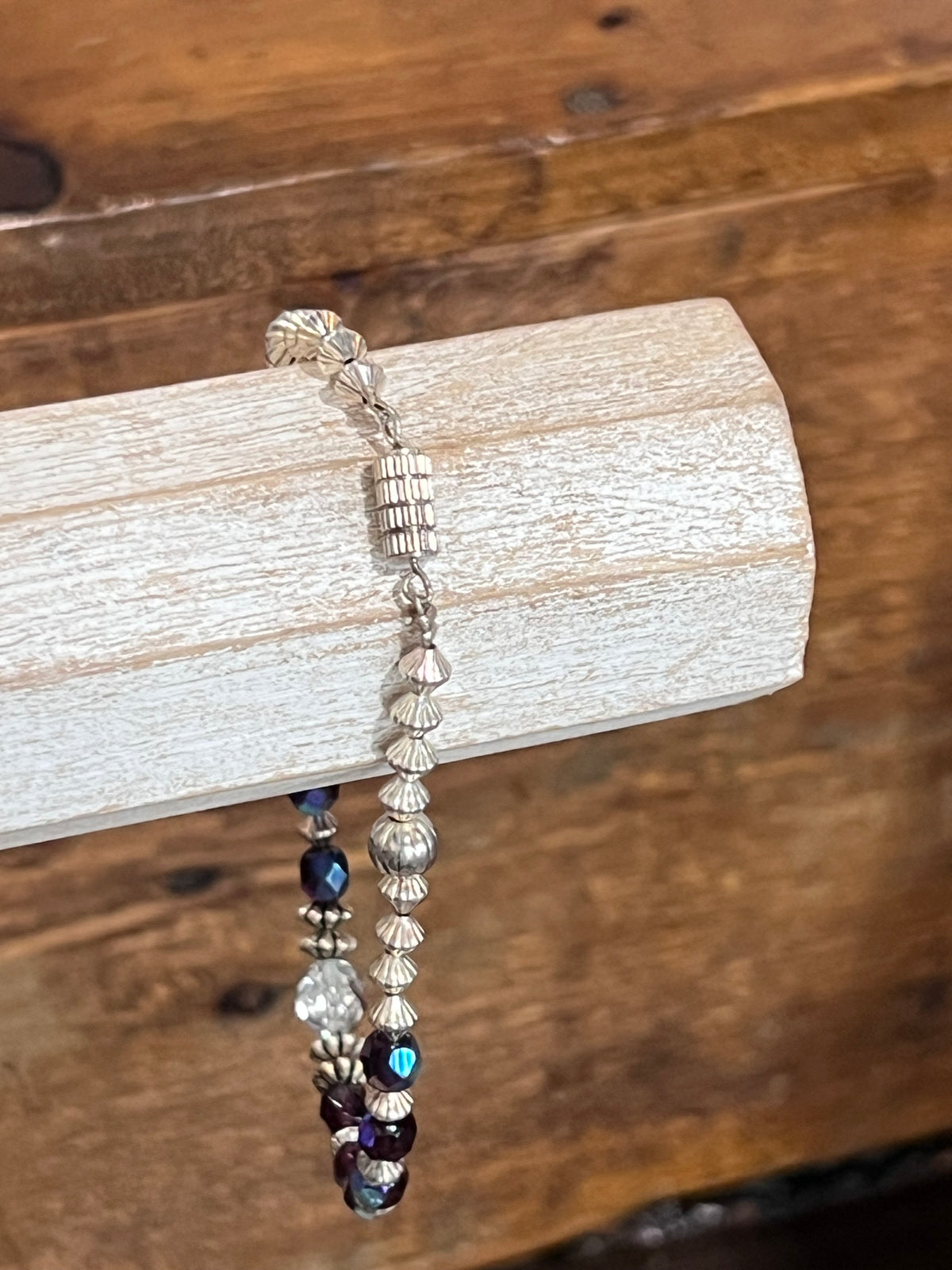 A beaded bracelet with silver-toned beads and dark purple iridescent beads, draped over a light-colored wooden block on a rustic wooden surface.