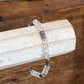 A beaded bracelet with clear faceted beads, iridescent blue-purple beads, and silver-toned spacer beads, displayed on a white wooden block over a wooden table.