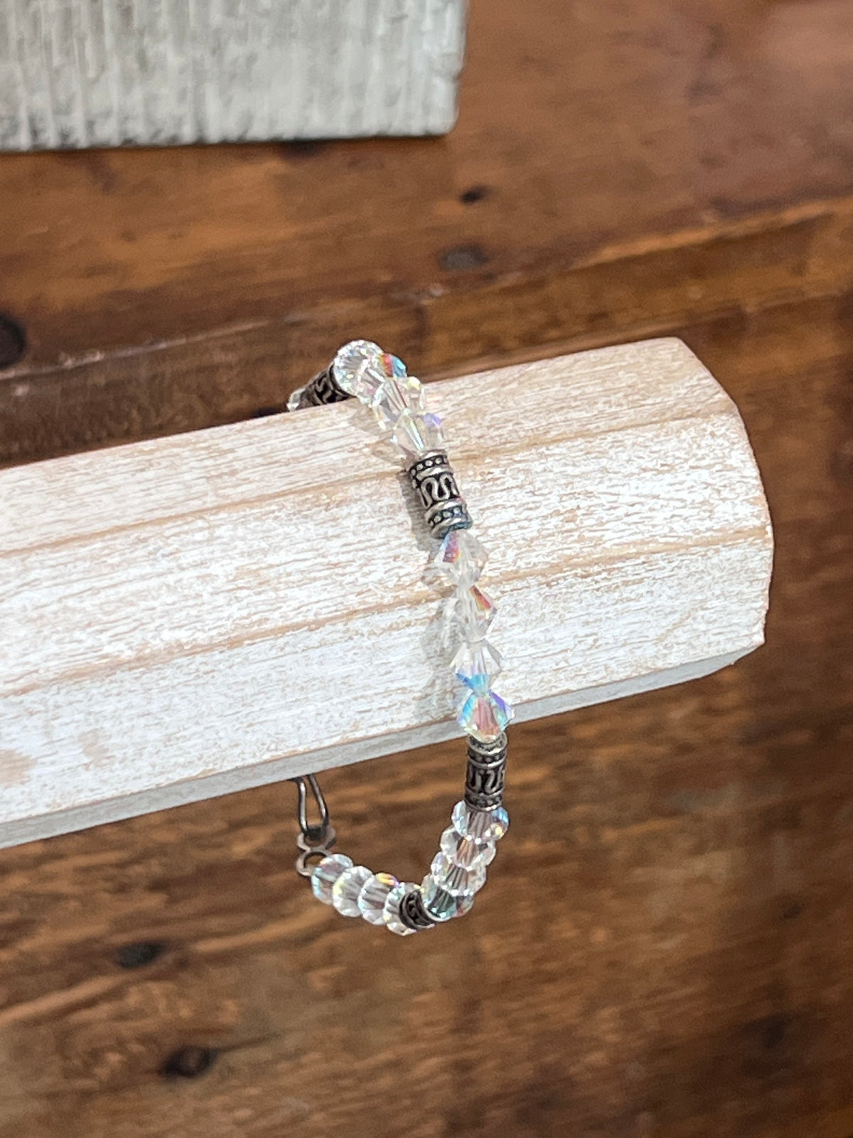 A beaded bracelet with clear faceted beads, iridescent blue-purple beads, and silver-toned spacer beads, displayed on a white wooden block over a wooden table.