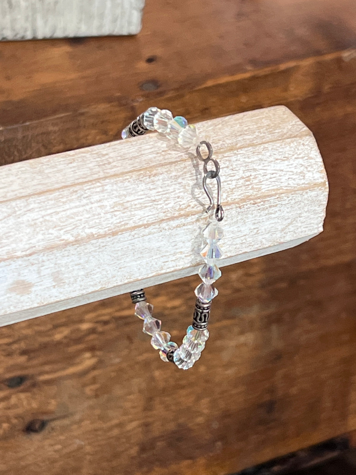 A beaded bracelet with faceted clear and iridescent beads, dark spacer beads, and a silver clasp, displayed on a light wood block over a wooden surface.