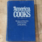 A vintage hardcover cookbook titled 'America COOKS' with a blue fabric cover and white lettering, placed on a light-colored wooden surface.
