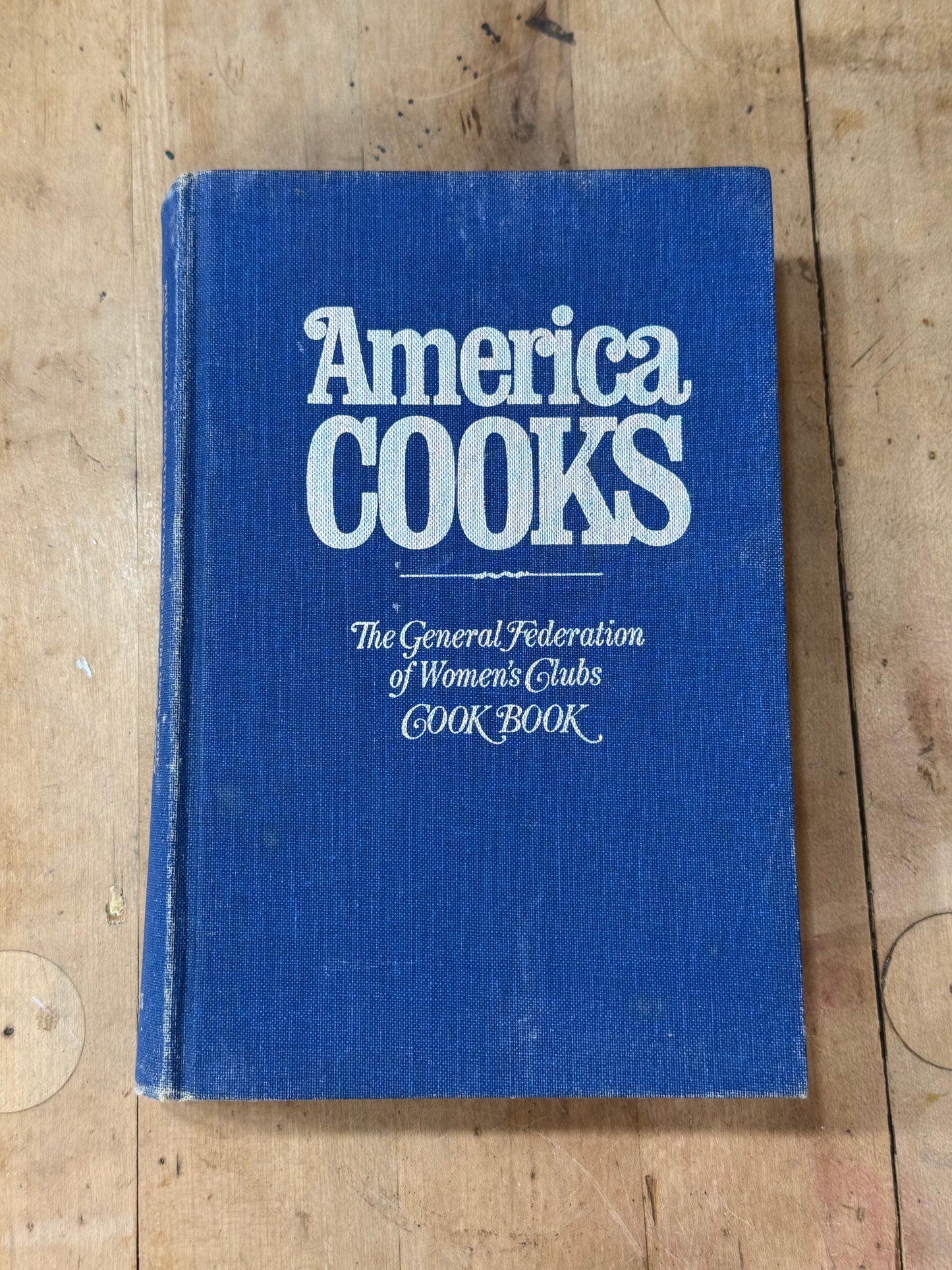 A vintage hardcover cookbook titled 'America COOKS' with a blue fabric cover and white lettering, placed on a light-colored wooden surface.