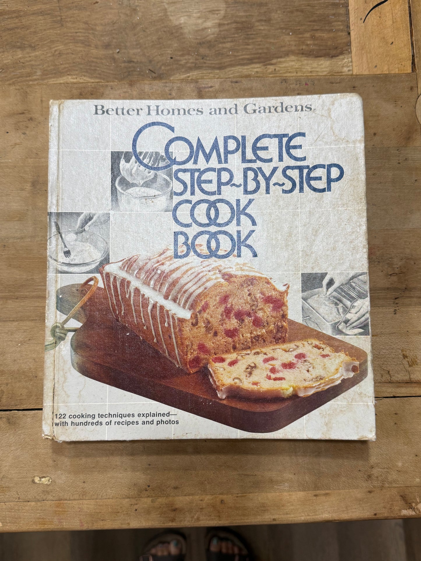 A vintage hardcover cookbook titled 'Complete Step-by-Step Cook Book' by Better Homes and Gardens, featuring a loaf of bread on the cover, placed on a wooden tabletop.