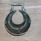 A vintage metal horseshoe-shaped pull tab or decorative hardware piece with a rectangular top loop, shown on a light brown wooden surface.