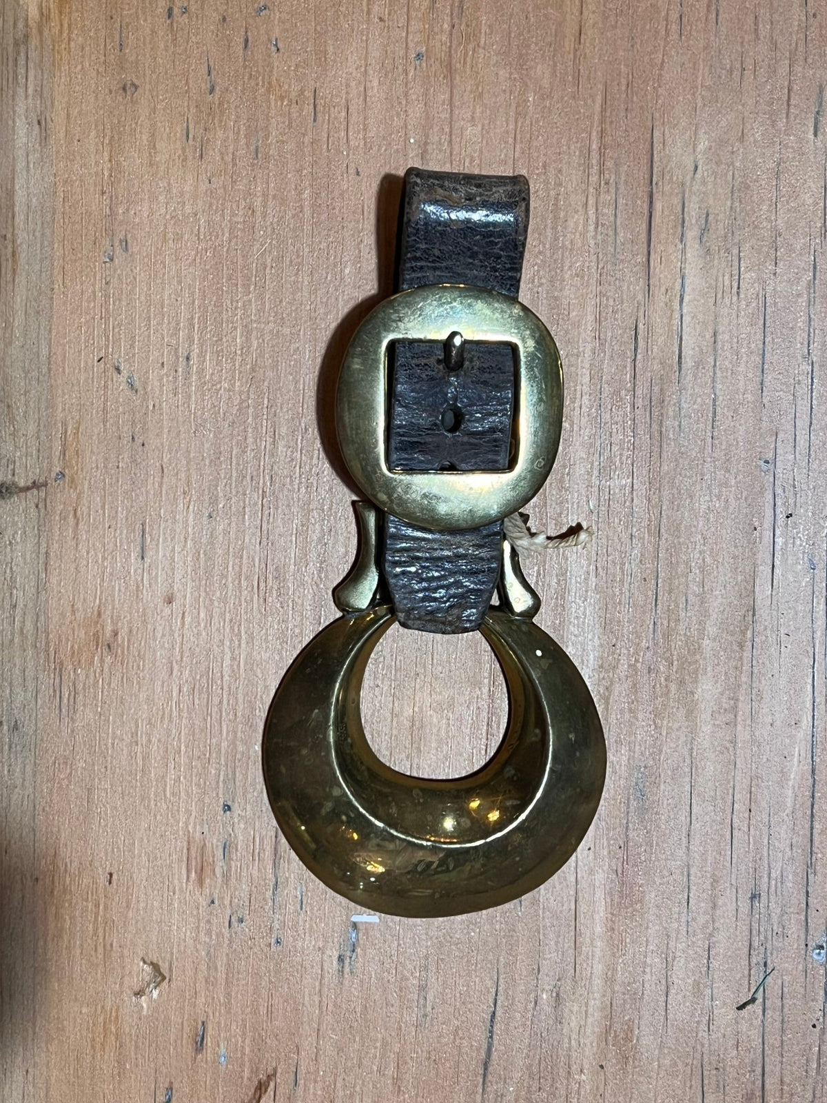 A vintage brass door bell with a dark leather strap and a large circular ring, displayed on a wooden surface.