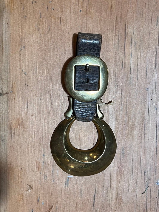 A vintage brass door bell with a dark leather strap and a large circular ring, displayed on a wooden surface.