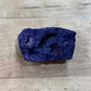 A raw, rough purple mineral specimen with a dark, textured surface, resting on a light-colored wooden surface.