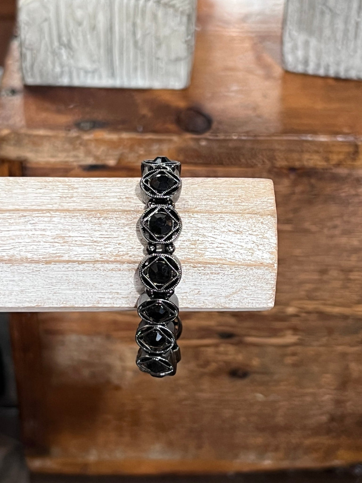 A black beaded stretch bracelet with faceted geometric beads, displayed on a light-colored wooden block over a rustic wooden surface.