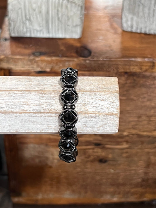 A black beaded stretch bracelet with faceted geometric beads, displayed on a light-colored wooden block over a rustic wooden surface.
