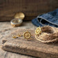 A pair of gold nautical ship's wheel stud earrings displayed on a rustic wooden board, alongside a matching gold compass and a coiled jute rope with a small gold charm, set against a background of denim and wood.