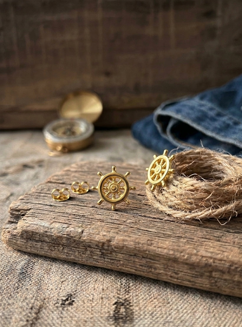 A pair of gold nautical ship's wheel stud earrings displayed on a rustic wooden board, alongside a matching gold compass and a coiled jute rope with a small gold charm, set against a background of denim and wood.
