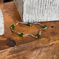 A silver wavy bracelet with five green faceted beads, displayed on a rustic wooden surface next to a textured gray block.
