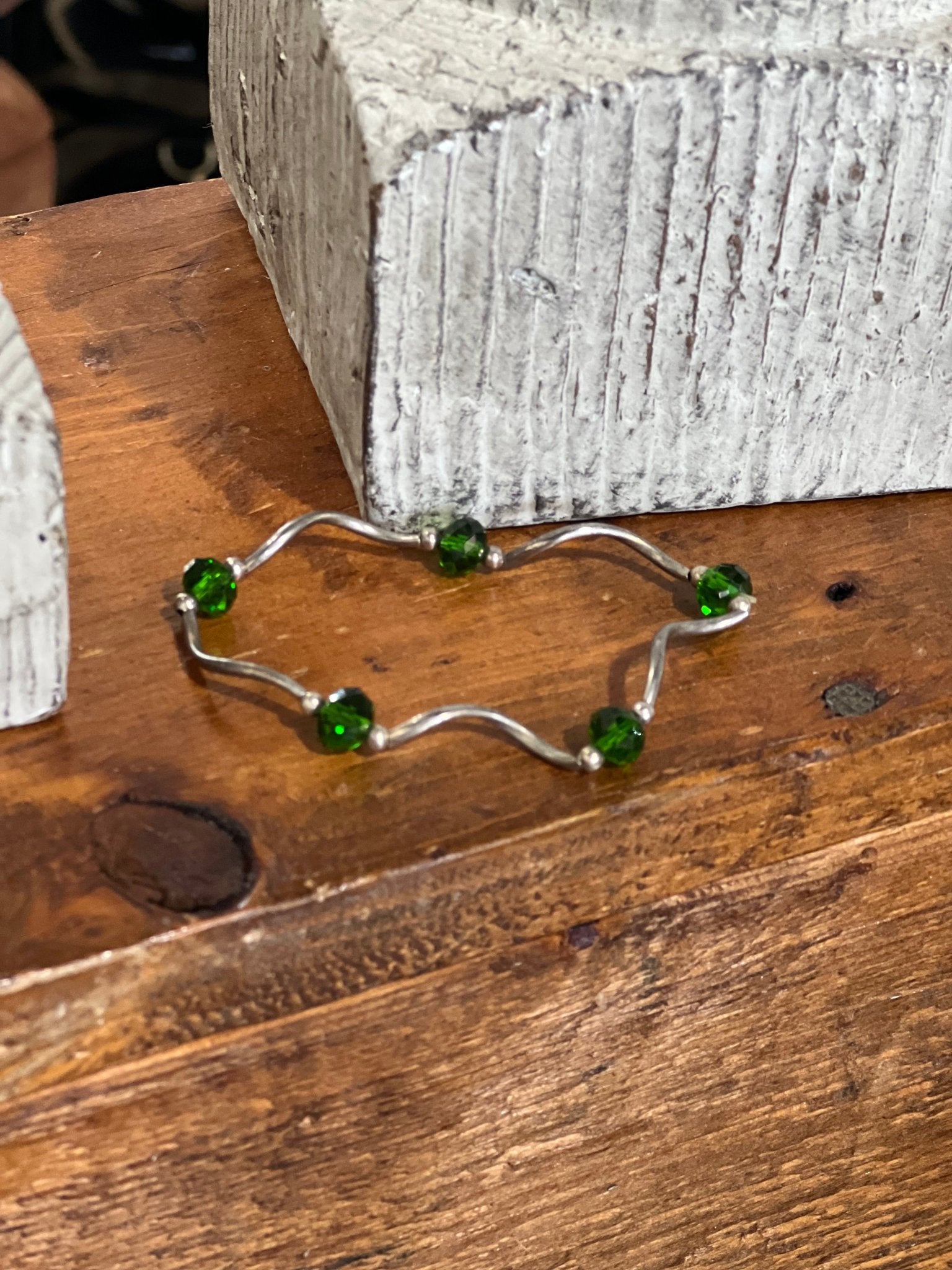 A silver wavy bracelet with five green faceted beads, displayed on a rustic wooden surface next to a textured gray block.