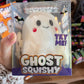 A packaged 'Crazy Fun Ghost Squishy' toy in clear plastic packaging, held in a hand. The product is a white ghost-shaped squishy with a purple bow, black eyes, and a red nose.