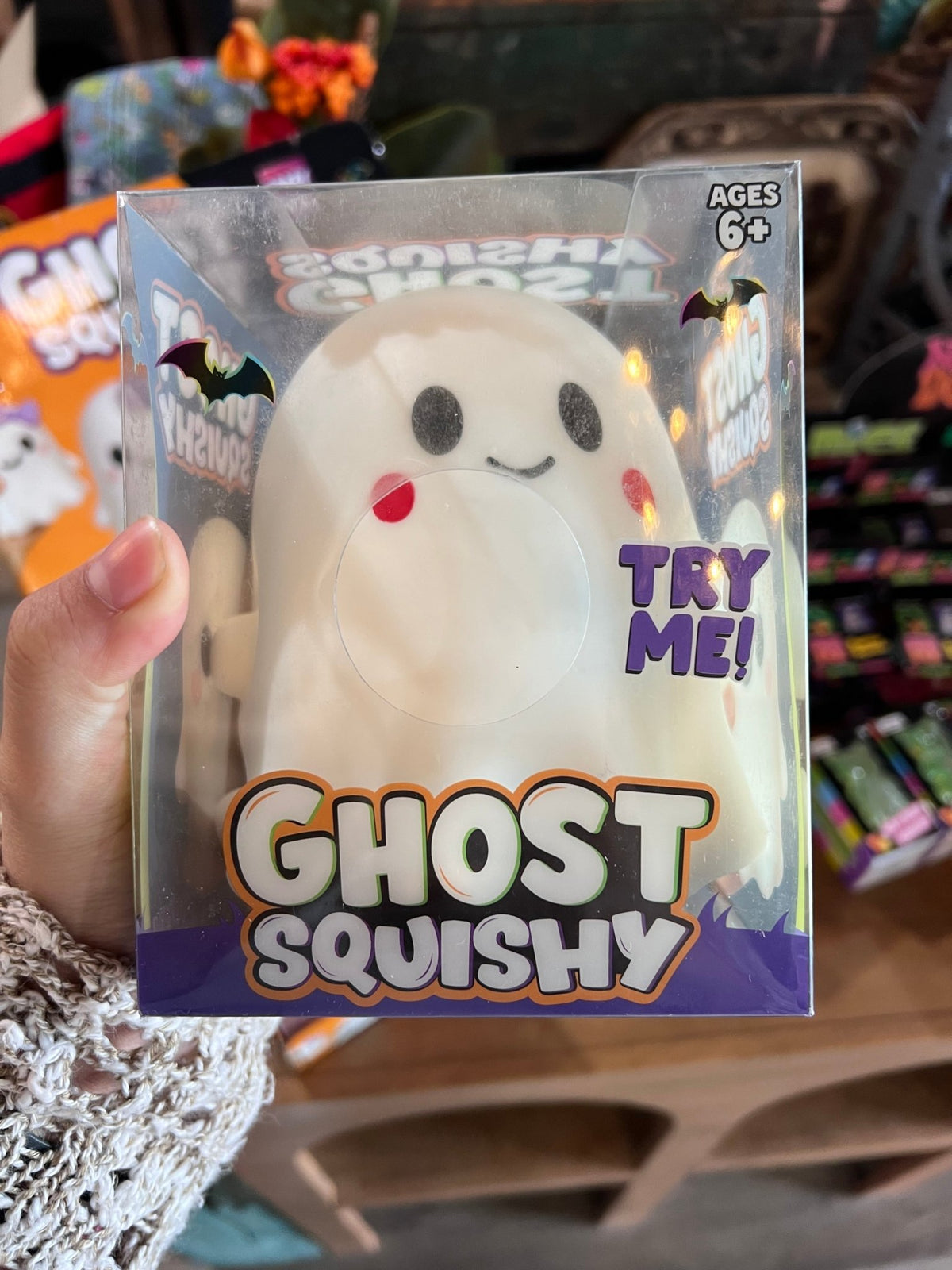 A packaged 'Crazy Fun Ghost Squishy' toy in clear plastic packaging, held in a hand. The product is a white ghost-shaped squishy with a purple bow, black eyes, and a red nose.