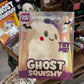 A packaged 'Crazy Fun Ghost Squishy' toy in clear plastic packaging, held in a hand. The product is a white ghost-shaped squishy with a purple bow, black eyes, and a red nose.