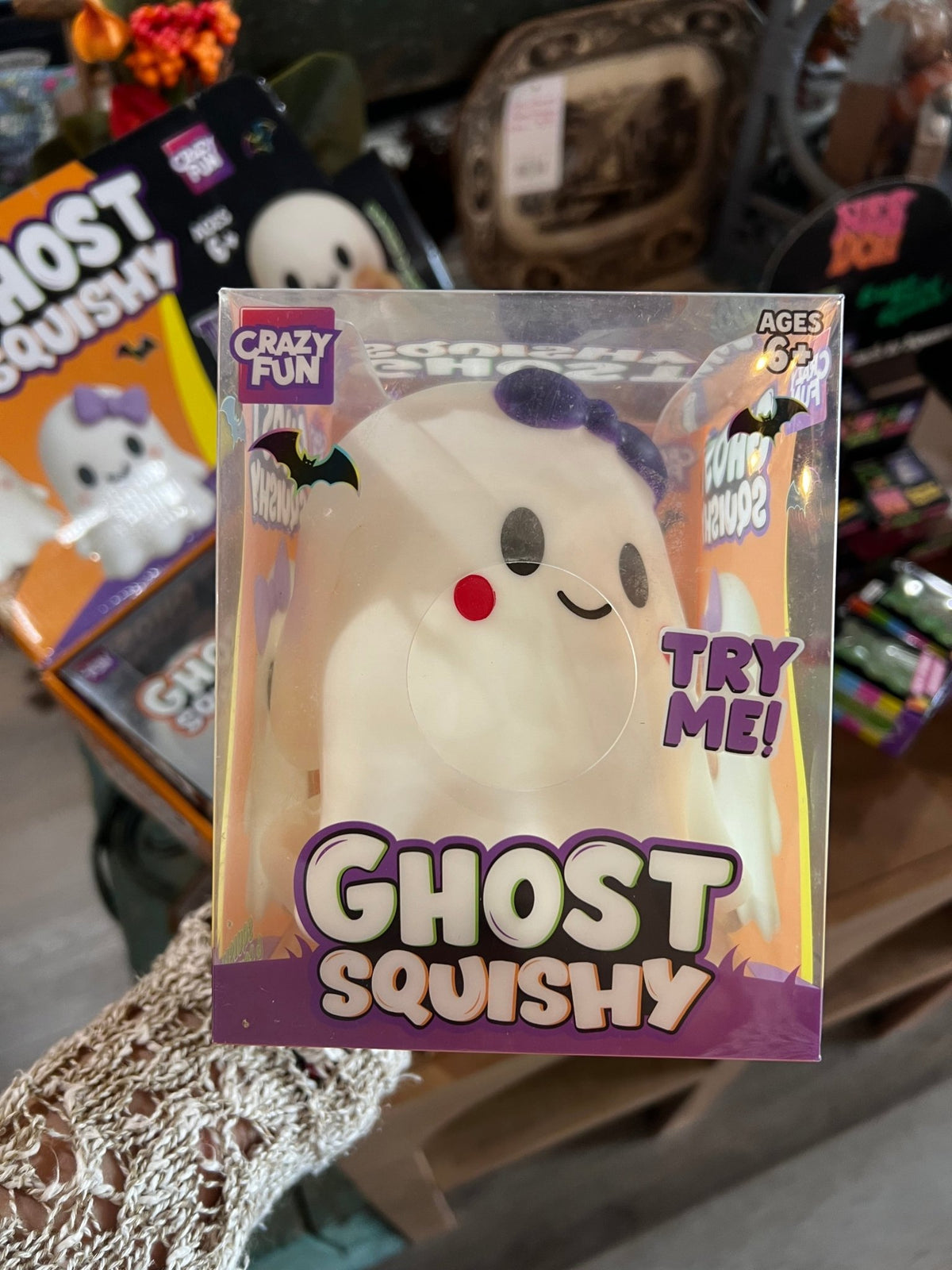 A packaged 'Crazy Fun Ghost Squishy' toy in clear plastic packaging, held in a hand. The product is a white ghost-shaped squishy with a purple bow, black eyes, and a red nose.