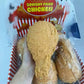 A hand holds a large, yellowish-brown, squishy fried chicken drumstick toy in front of its retail packaging box labeled 'Squishy Fried Chicken'.