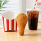 A golden-brown fried chicken drumstick stands upright on a light wood table next to a red and white striped paper fry container and a clear cup of iced cola with a red-and-white striped straw.