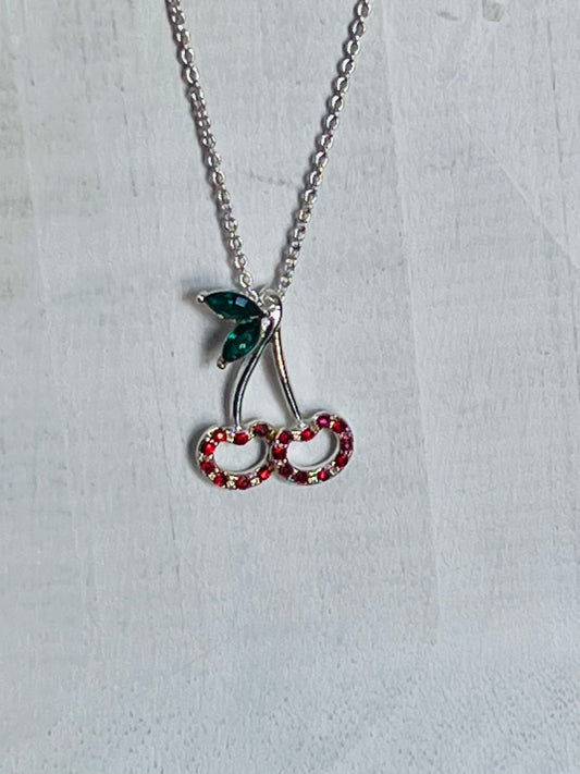 A silver-tone cherry pendant necklace with a green leaf and red rhinestone-studded cherries on a textured white background.