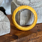 A twisted yellow bangle bracelet displayed on a rustic wooden surface in front of textured gray stone props.