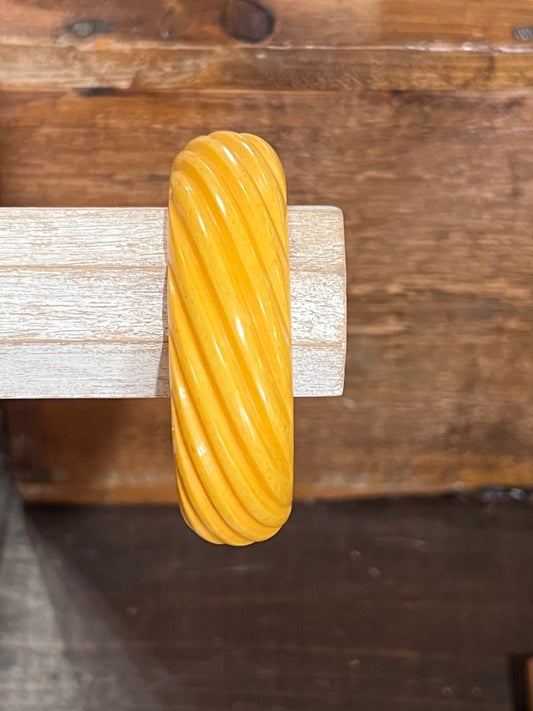 A thick, glossy yellow twisted bangle bracelet displayed on a light-colored wooden stand against a rustic dark wood background.