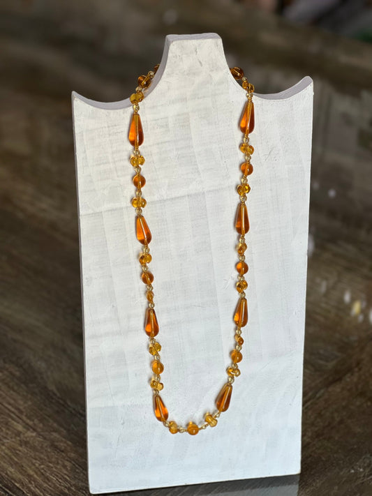 An amber glass bead necklace with teardrop-shaped beads and small round beads, displayed on a white fabric-covered jewelry stand against a dark wooden background.