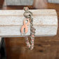 A silver-toned chain bracelet with a pink enamel ribbon charm, displayed hanging from a rustic white wooden block over a dark wood surface.
