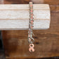 A silver-toned chain bracelet with a peach-pink link accent and a dangling pink awareness ribbon charm, displayed on a rustic wooden block.