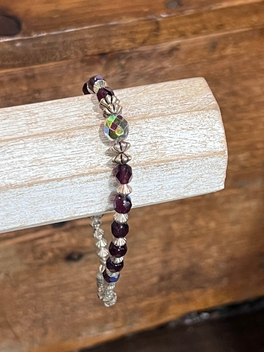 A beaded stretch bracelet with dark purple faceted beads, silver-toned textured beads, and a central iridescent green bead, displayed on a light wood block over a rustic wooden background.