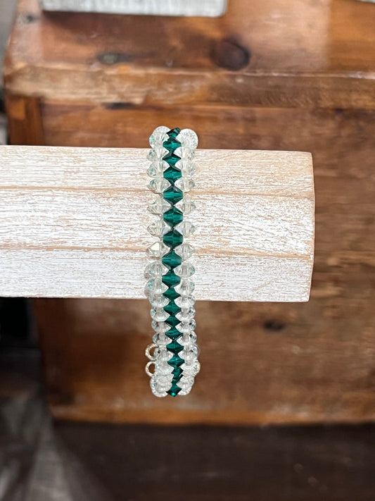 A beaded stretch bracelet with alternating dark green and clear faceted beads, displayed on a rustic white-painted wooden dowel with a wooden box in the background.