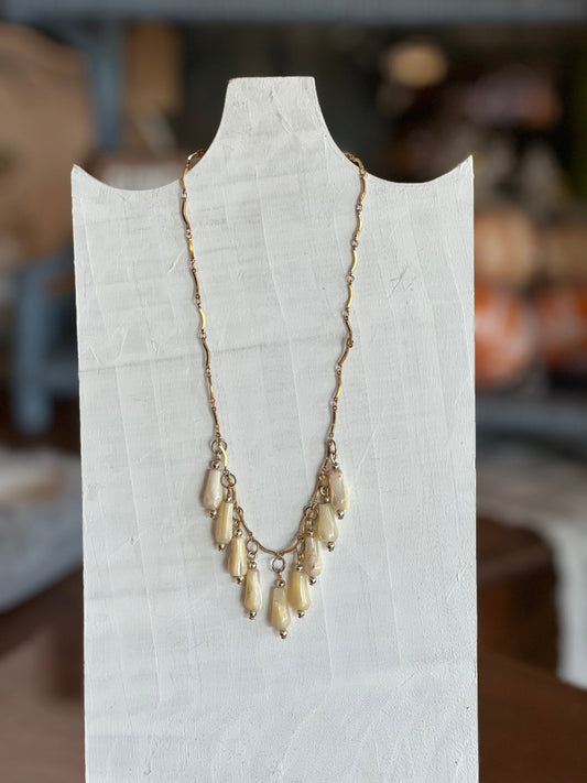 A gold-toned necklace with multiple cream-colored teardrop beads, displayed on a white fabric mannequin bust against a blurred indoor background.