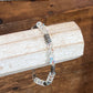 A beaded bracelet with clear faceted beads, iridescent blue-purple beads, and silver-toned spacer beads, displayed on a white wooden block over a wooden table.