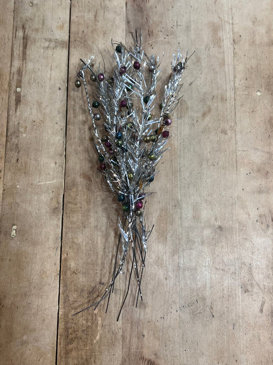 A decorative silver tinsel pine branch ornament with small multicolored beads, placed on a rustic wooden surface.
