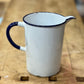 A vintage white enamel pitcher with a dark blue rim, handle, and spout, sitting on a rustic wooden workbench.