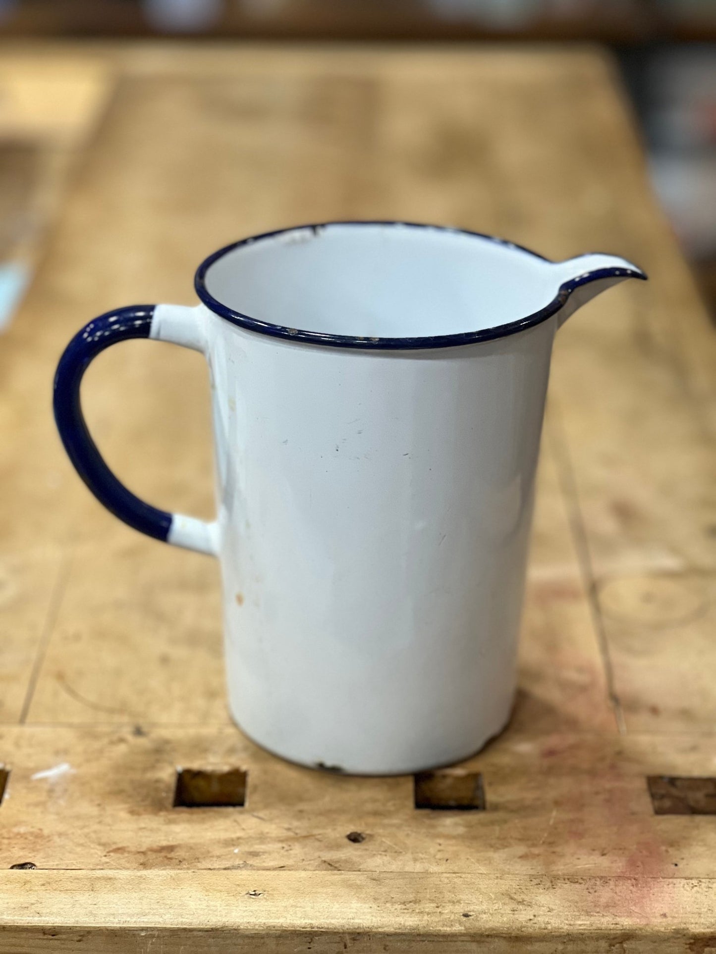 A vintage white enamel pitcher with a dark blue rim, handle, and spout, sitting on a rustic wooden workbench.