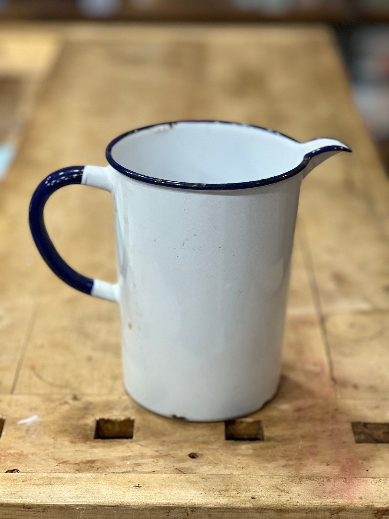 A vintage white enamel pitcher with a dark blue rim, handle, and spout, sitting on a rustic wooden workbench.