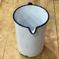 A white enamel pitcher with a dark blue rim and handle, shown from a top-down view on a wooden workbench.